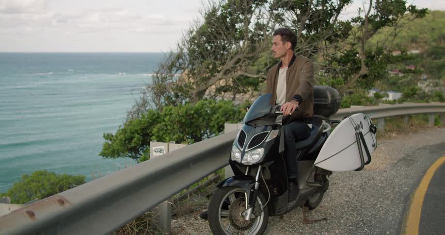 A man sits on his scooter on the side of the road by the water