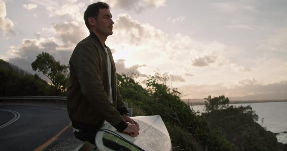 Young man waxing a surf board on a road near the coast at sunset