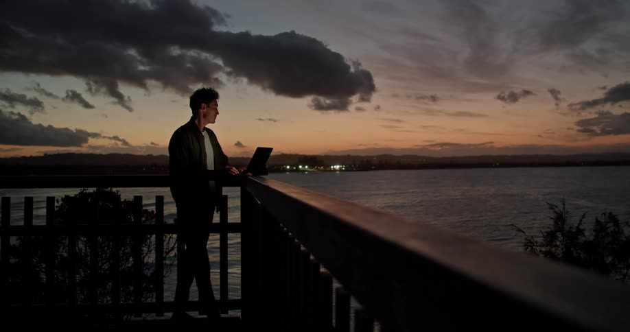 Man stand near rail typing on computer near ocean under fading afterglow