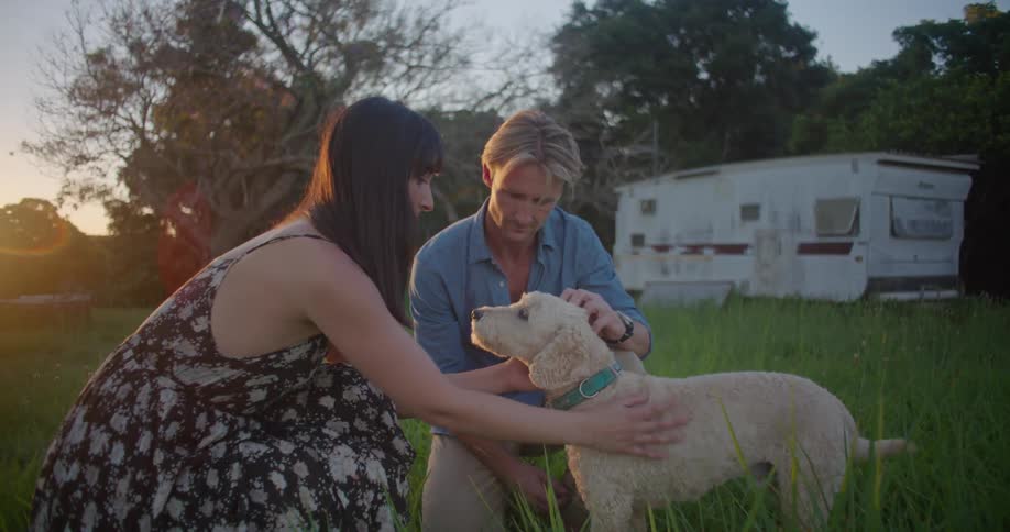 A man and woman pet a small dog while kneeling in a field during the day