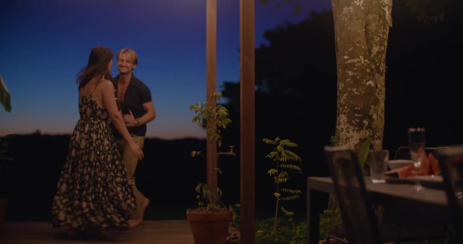 A couple are dancing on a patio at a house in a rural location at dusk