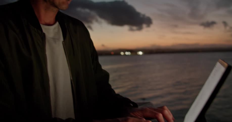 Man wearing white t shirt and black jacket types on tablet keyboard on the beach next to sea at dusk