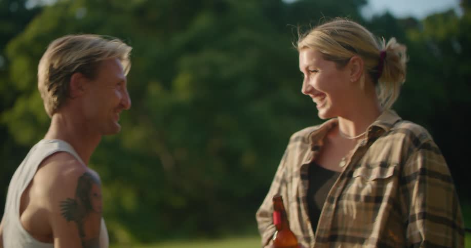 A woman and man holding beer bottles talk and laugh together while sitting outdoors during the day