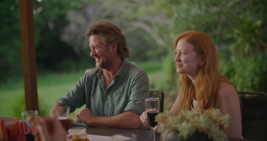 A man and woman sitting outside on a patio laugh as they eat and drink wine and beer with another person