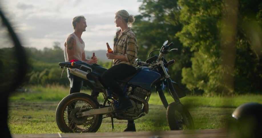 Woman sits on motorcycle in field near standing man talking before they toast with beer bottles