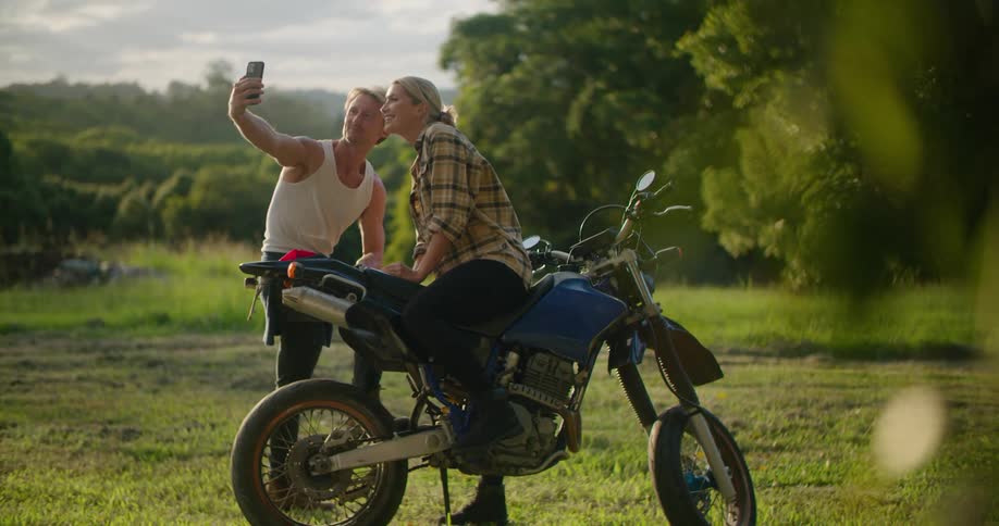 Blonde man with undershirt takes selfie with smartphone with blonde woman sitting on blue motorcycle in the field next to trees