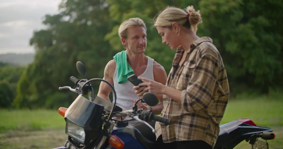 Young couple standing near a motorcycle parked in the countryside checking a phone