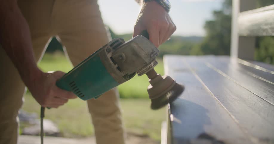 A man uses a sander to grind down the edge of a wooden bench