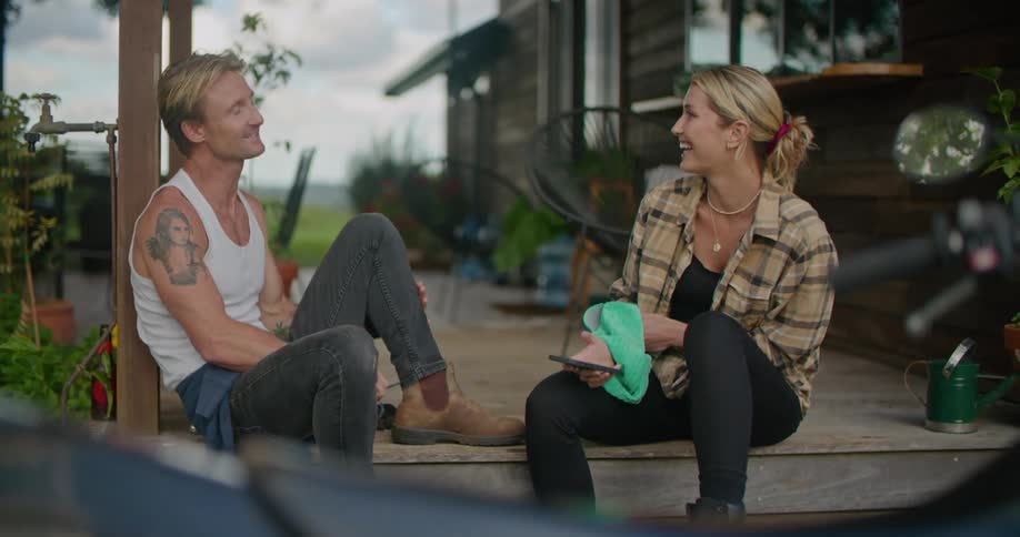A woman laughs while chatting with a man as they sit together on the porch of a house
