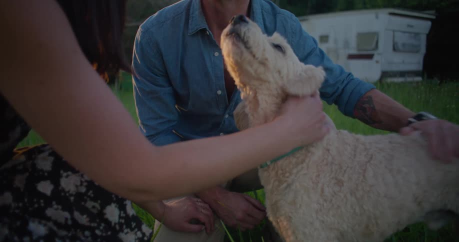 Blonde man with blue shirt brown pants tattoo and wristwatch pets brown dog with woman with brown wavy hair and dress in the countryside in front of trailer next to forest at sunset
