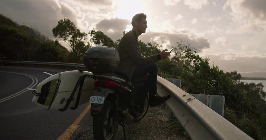 A man uses his smartphone casually while sitting atop a motorcycle parked on the shoulder of a winding mountain road