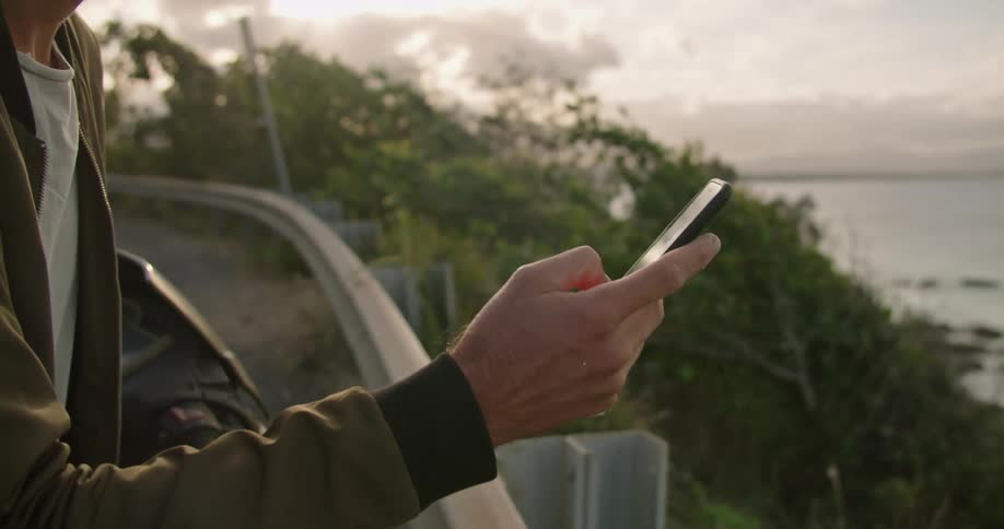 Man wearing jacket stands near rail on road using mobile phone while looking out over ocean
