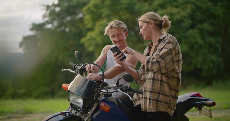 Couple sitting on a parked motorcycle in the countryside checking a phone at sunset