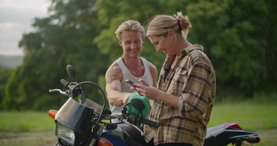 A woman sits on the motorcycle showing a man her cell phone