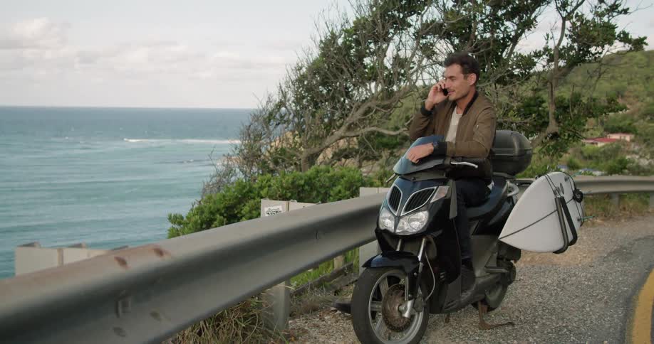 A man talks on his smartphone while sitting on a motorcycle parked in the shoulder of a road overlooking the ocean