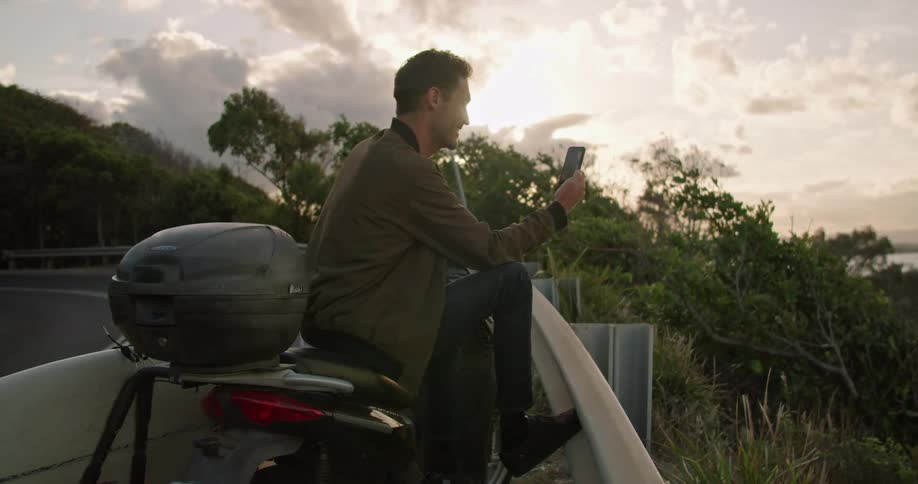 A man leans against his motorcycle and laughs and waves at his cell phone as he video chats with someone