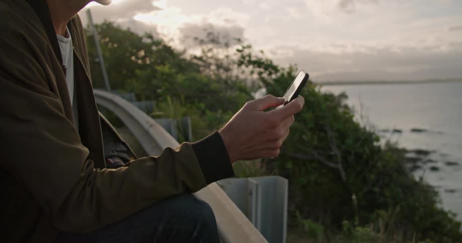 Man standing on side of road overlooking shore texts as car passes behind him