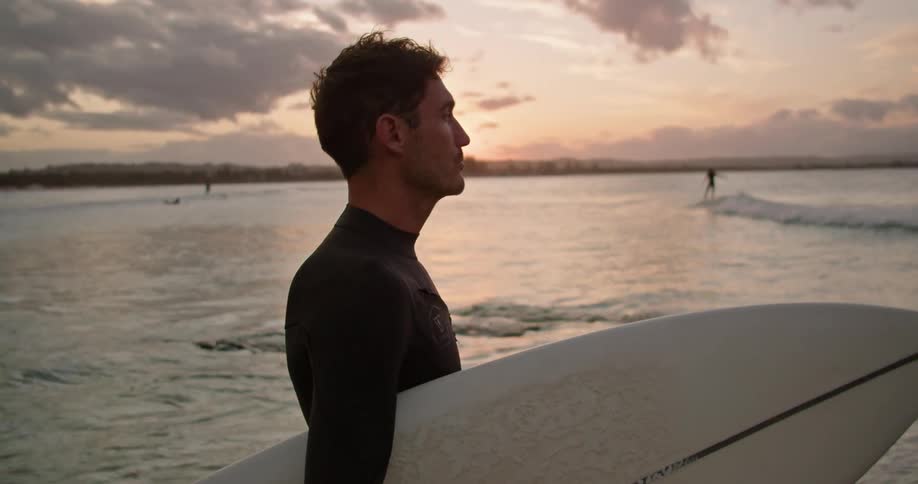 A man wearing a wetsuit and holding a surfboard is standing on a beach as the sun is setting