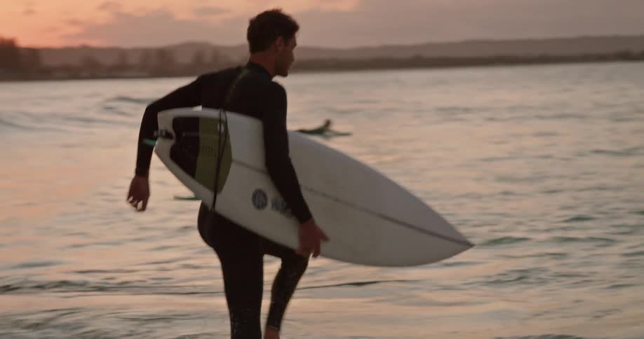 A man wearing a wetsuit and holding a surfboard is standing on a beach and walking into the water
