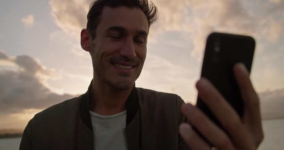 A man waves and blows a kiss while talking on his phone with the ocean behind him