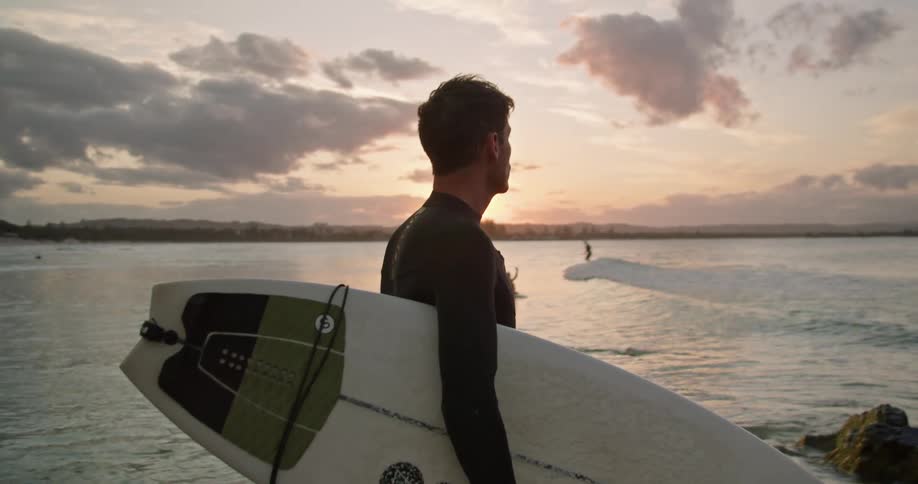 Male surfer with wetsuit holds surfboard and looks at sea at sunset on the beach