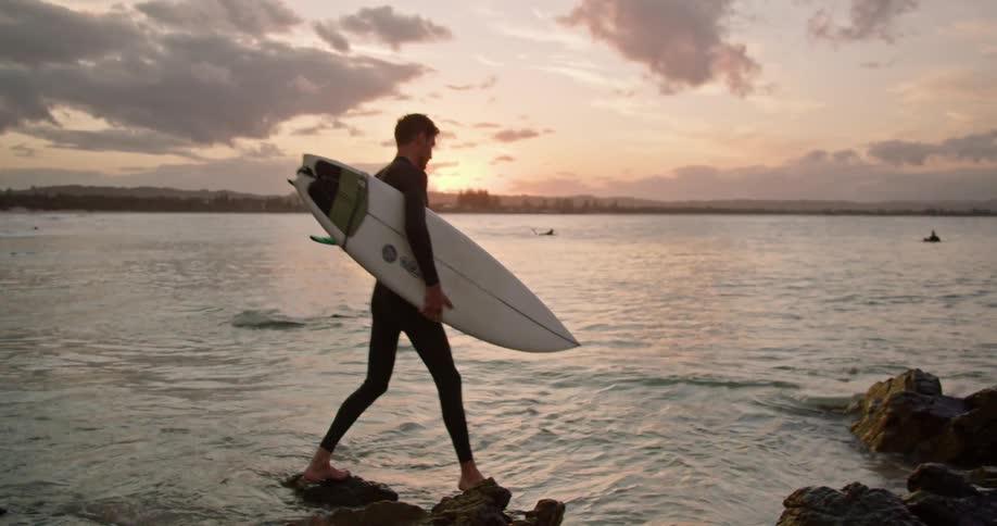 Surfer carrying surfboard moves wades into water near beach as sun turns horizon pink