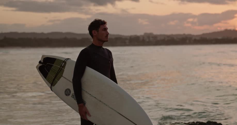Man stands holding surfboard under red sky as another surfer paddles out lying on his board