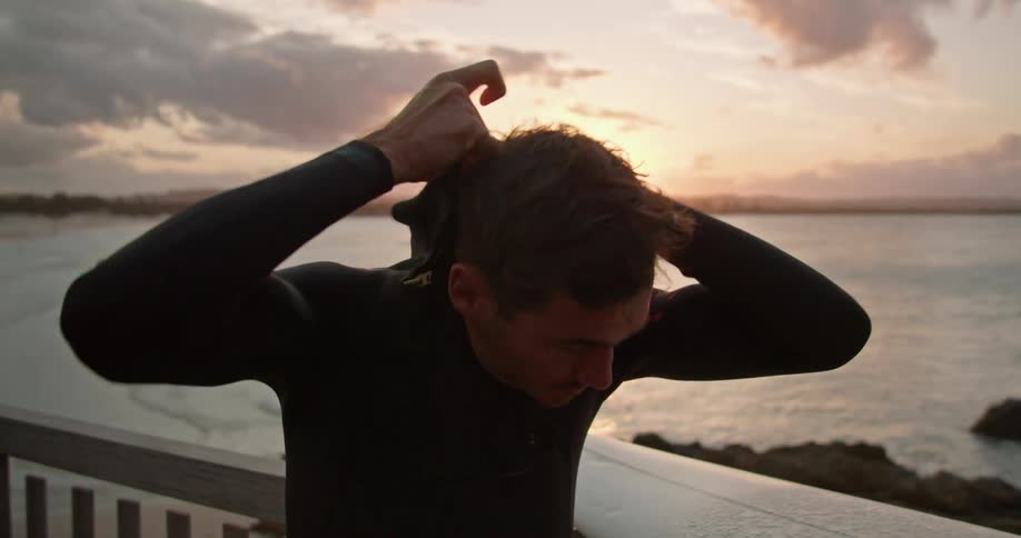 A surfer standing on a bridge above a beach puts on a wet suit before carrying his surfboard away out of frame