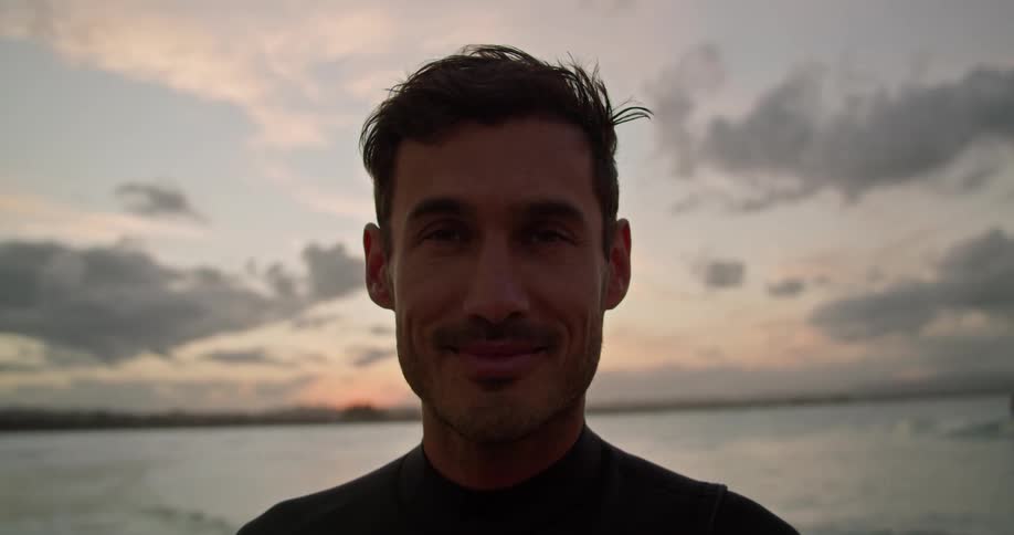 Male surfer with wetsuit looks at camera on the beach next to sea at dusk
