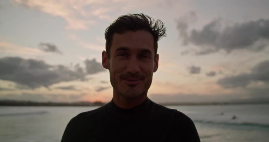 A man in a wetsuit smiles as he stands near the ocean