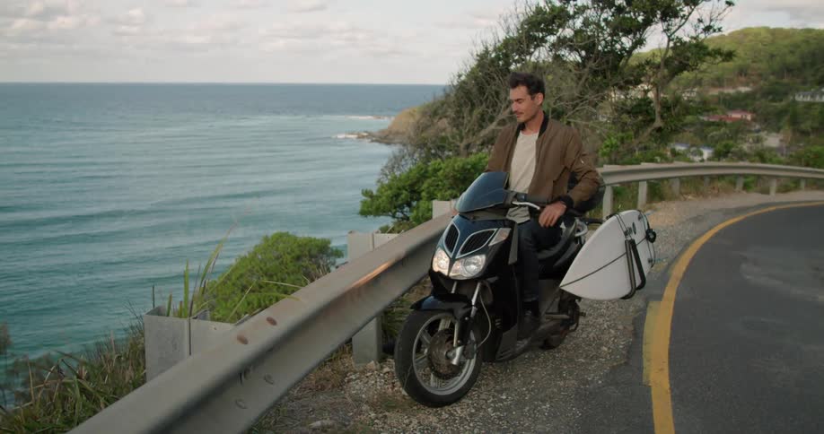Man sitting over a motorcycle on a road next to the ocean at sunset