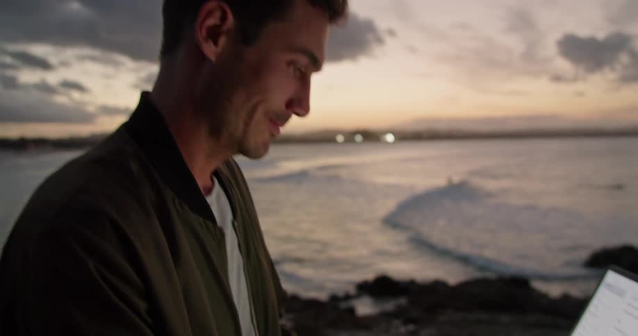 A man stands on a balcony on the shore and types on a laptop