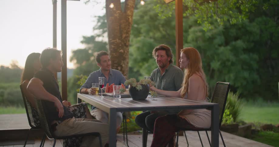A group of friends sitting at table drinks alcohol and talks to each other on terrace in the countryside