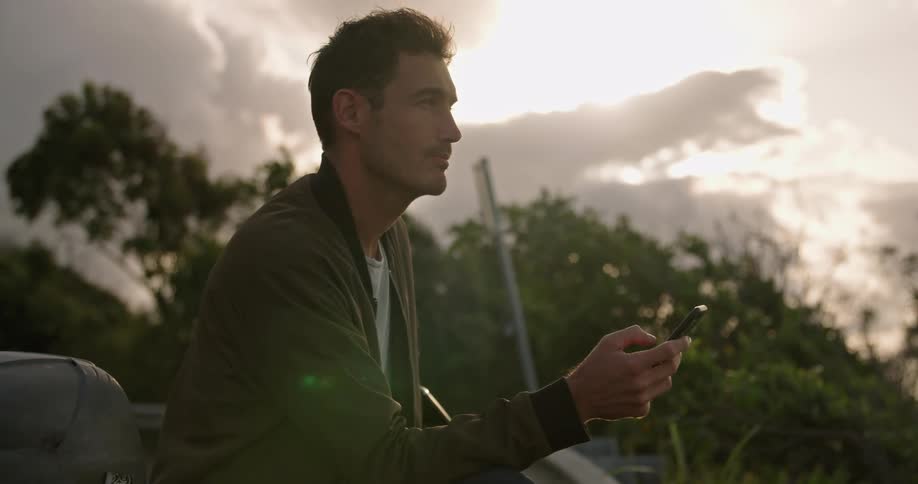 A man is sitting outside under a cloudy sky with trees in the distance making a call on his cell phone