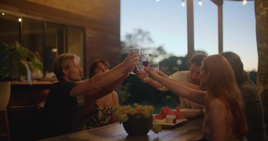 A group of people sitting around a wooden dining table raise their glasses in a toast
