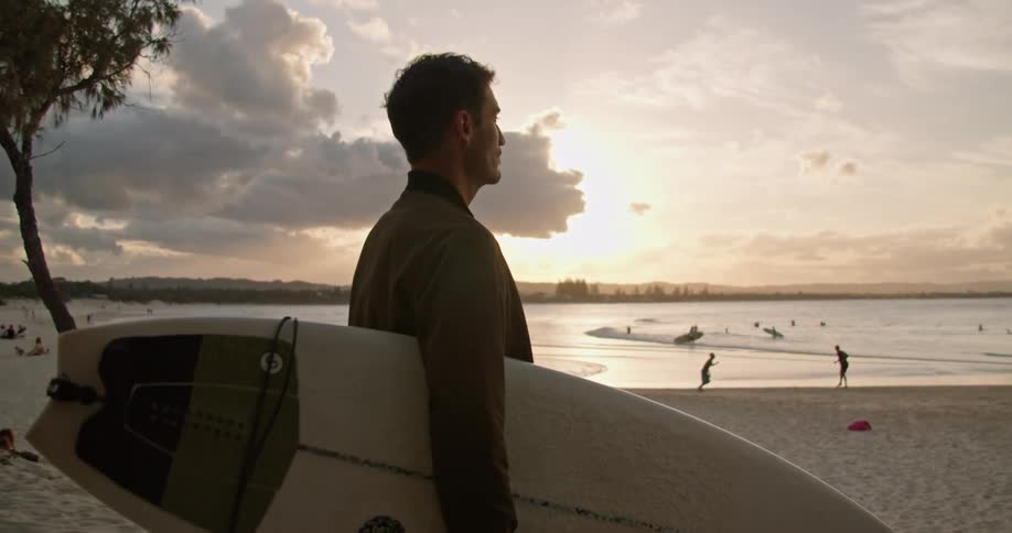 A man holding a surf boar stands on the beach and looks around as children play ball and people play in the water and then he walks towards the water