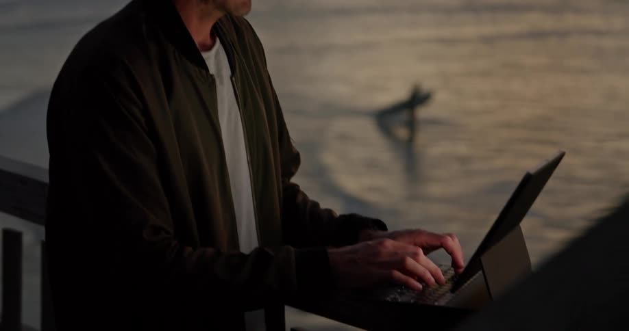 Man sits near rail typing while surfers move through ocean in background