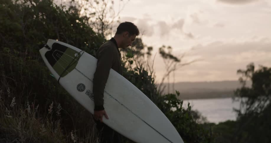 Young man carrying a surfboard climbs down a hill to get to a path near the coast at sunset