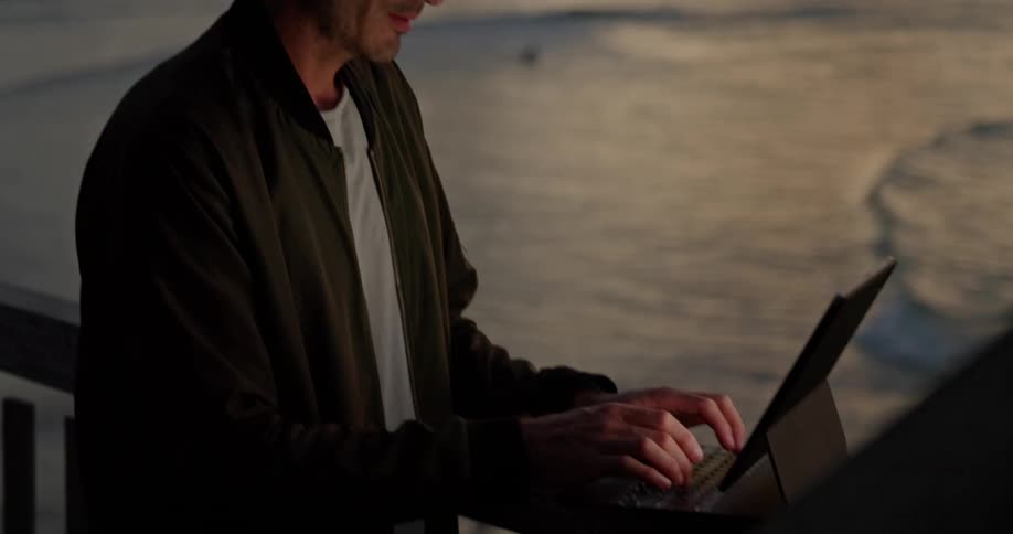 A man types on a laptop propped up on a railing overlooking the ocean