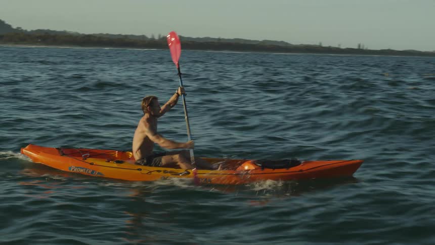 Young shirtless man rowing on a red kayak on the ocean at sunset