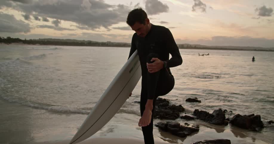 Young man wearing a black wet suit flexes while holding a surfboard near the beach at sunset