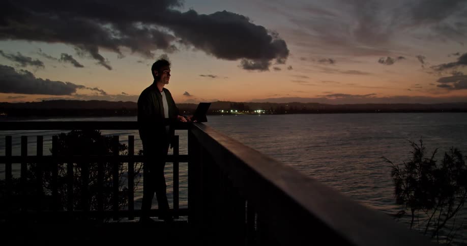Man stands typing on computer sitting on banister overlooking water with town lights visible under red sky on opposite shore