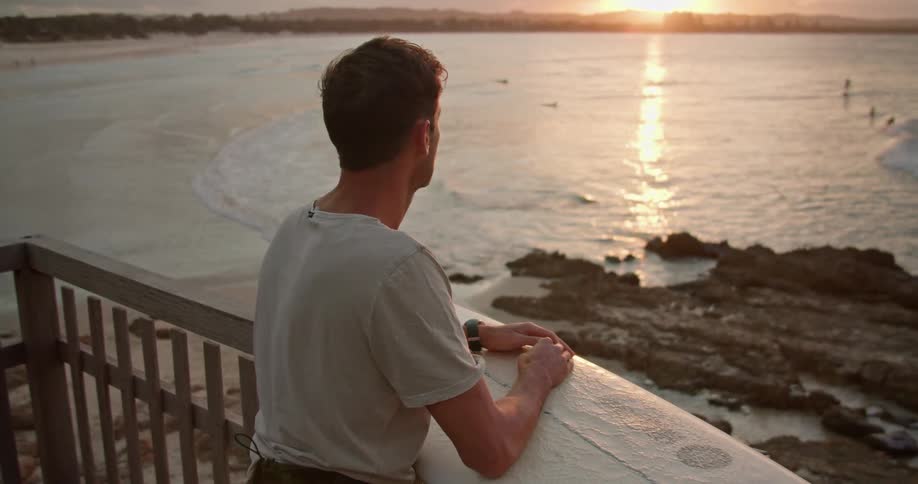 A man leans on his surfboard propped on his balcony as he watches people surfing in the ocean