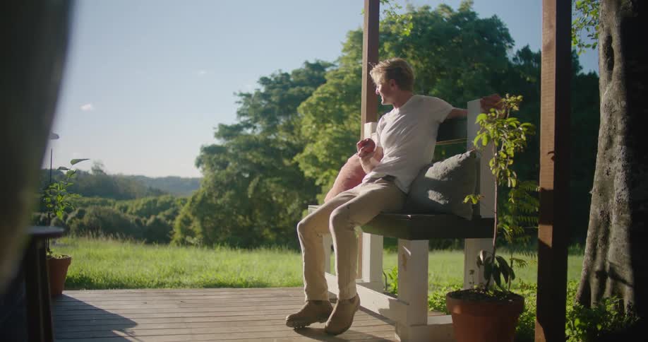 A man places a cushion on a wooden bench outdoors before sitting down and taking a bite from an apple