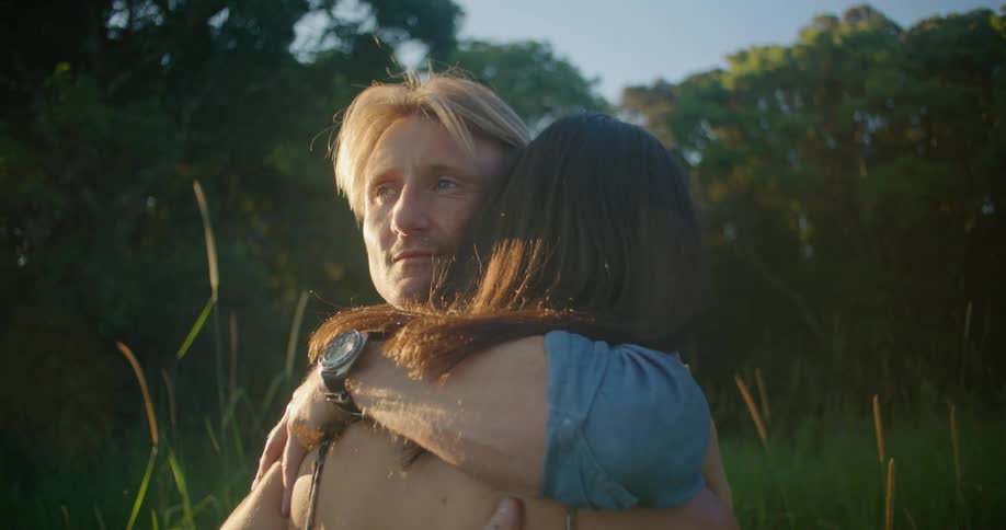 Blonde man with blue shirt and wristwatch cuddles woman with brown wavy hair and dress in the countryside next to trees