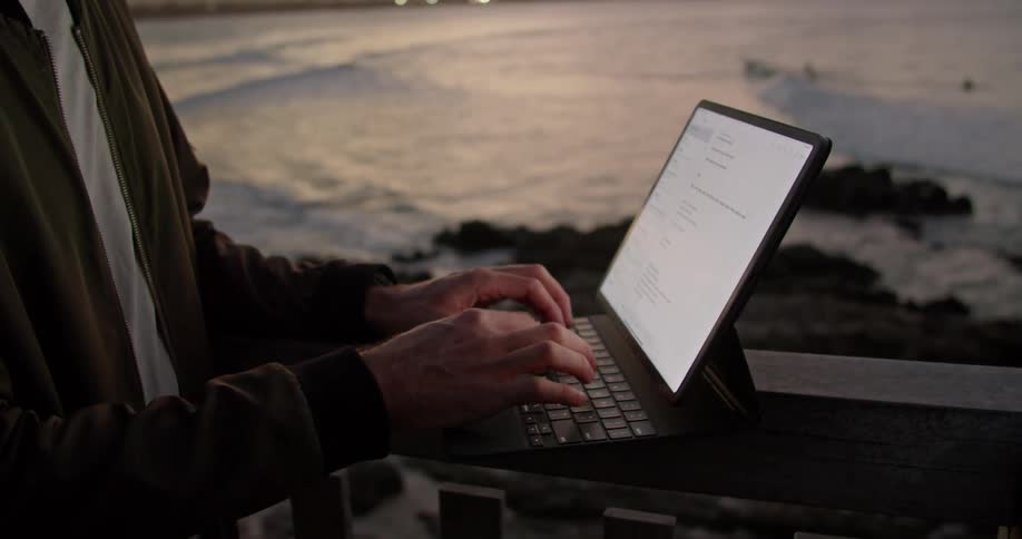 A man is standing near a railing overlooking the water and typing on a laptop computer