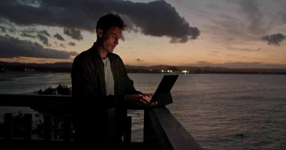 A man is standing near a railing overlooking the water and typing on a laptop computer