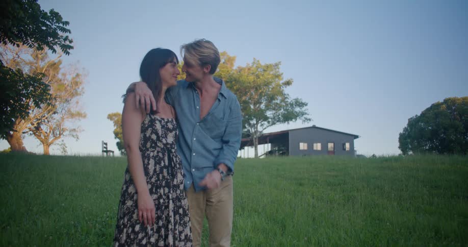 Blonde man with blue shirt and brown pants holds woman wearing dress in the countryside kissing each other