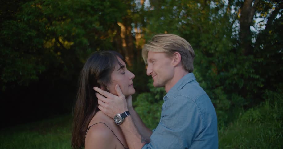 Blonde man with blue shirt kisses woman with brown wavy hair and dress lovingly looking into each others eyes in the countryside next to trees