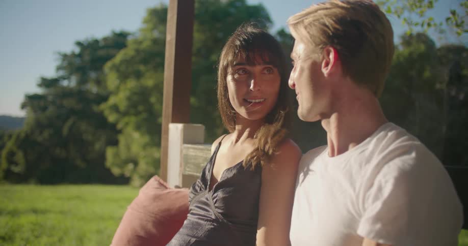 Couple sit on bench outdoors on sunny day looking around and kissing